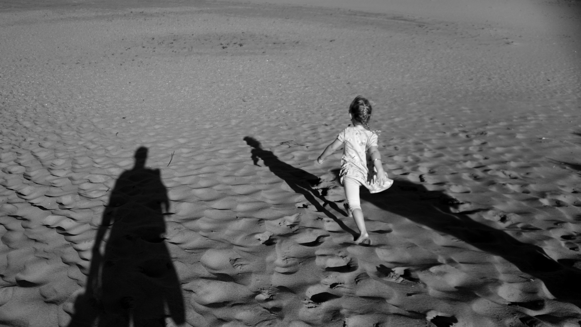 Kid running away from the camera on an empty beach. Shadows of her parents stretching in the sun